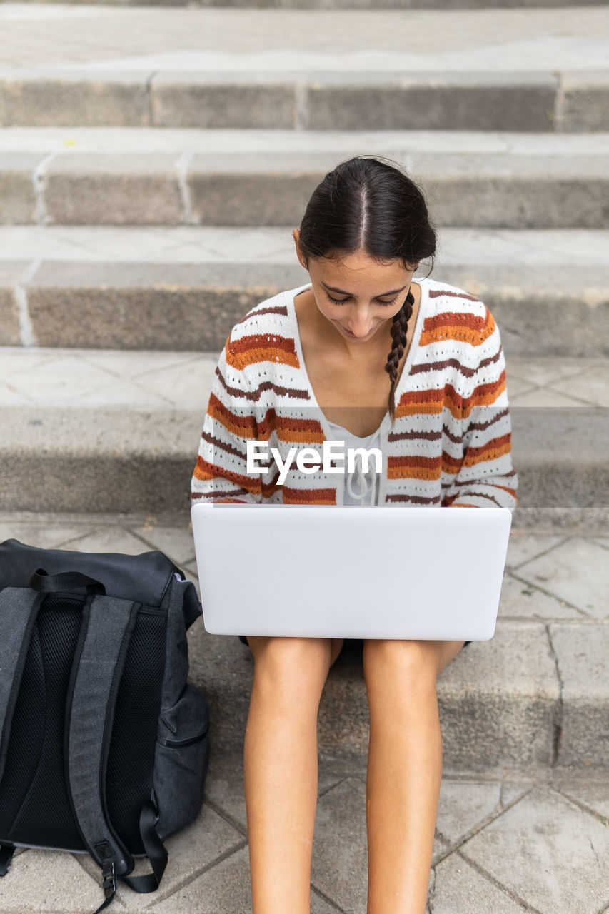 High angle of young female student browsing netbook and doing homework while sitting on concrete steps near backpack in city