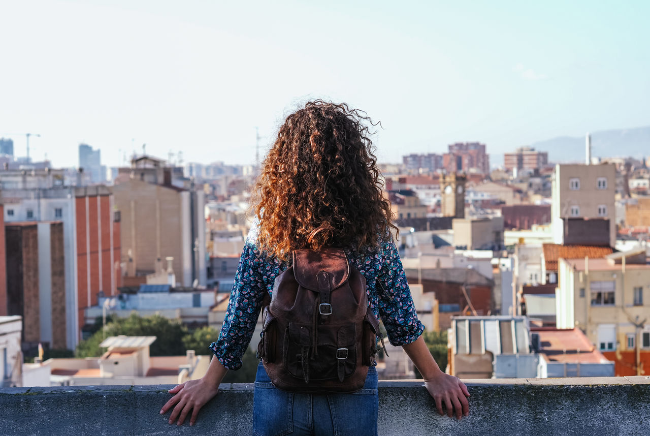 Back view of tranquil female with long curly hair standing on roof of building against cityscape while enjoying summer weekend