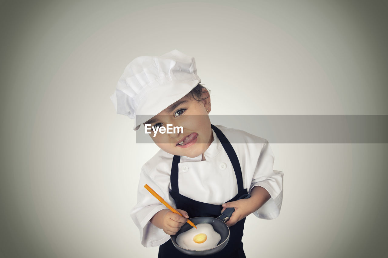 Portrait of girl in chef uniform preparing fried egg against white background