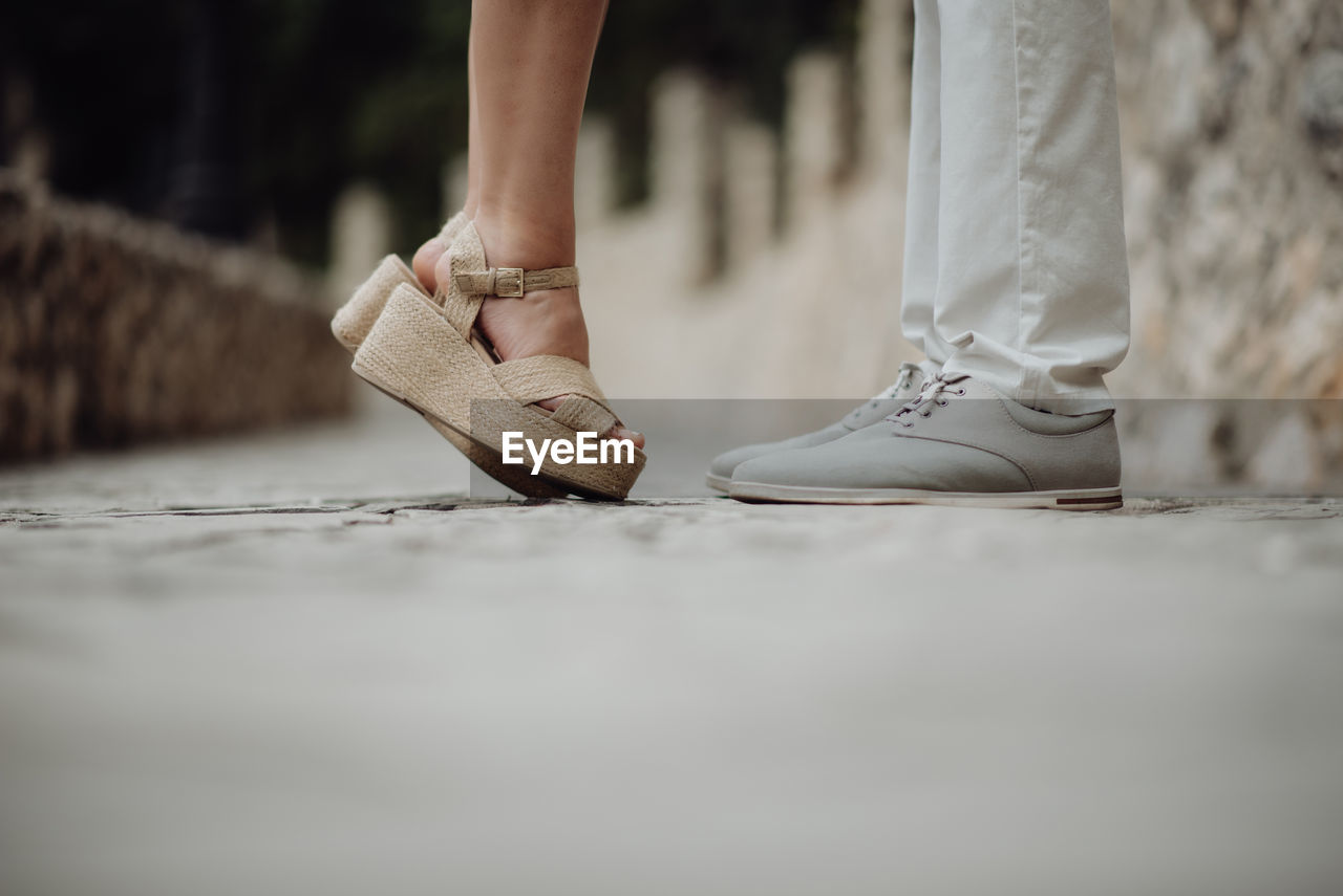 Crop side view of woman in sandals standing on tiptoe in front of casual man on pebbled pavement