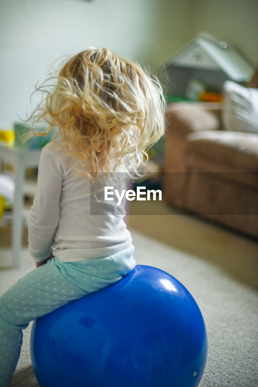 Rear view of girl sitting on fitness ball in living room at home