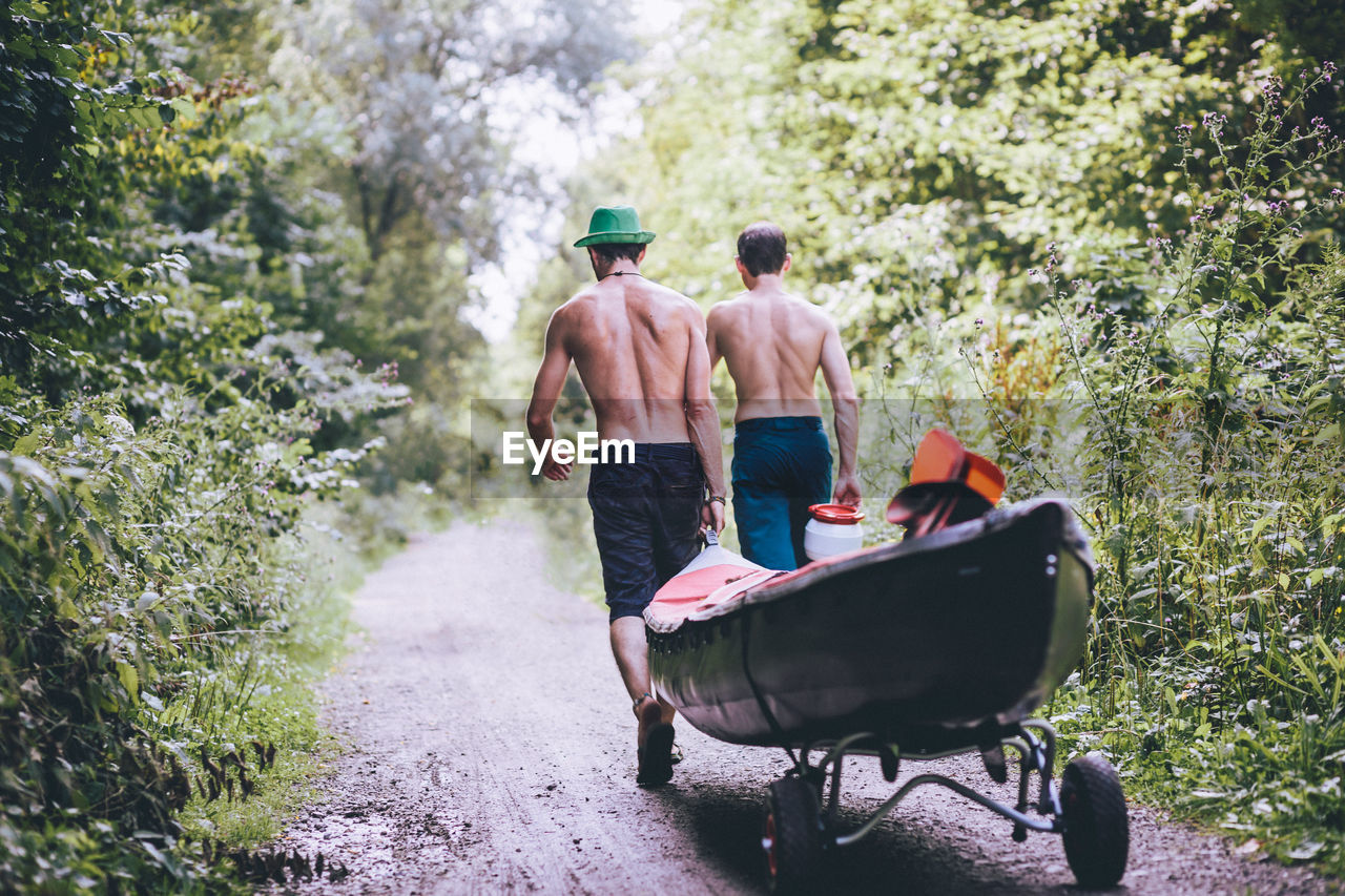 Rear view of shirtless friends carrying boat in forest