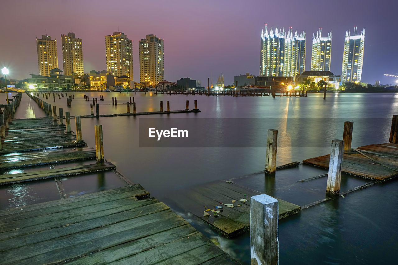 Pier on river by illuminated buildings against sky at night