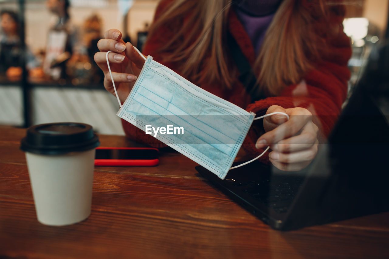 Midsection of woman holding mask while using laptop in cafe seen through window