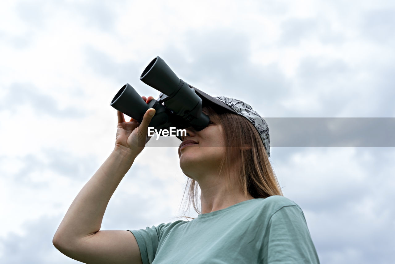 Young blonde woman bird watcher looking through binoculars at cloudy sky ornithological research 