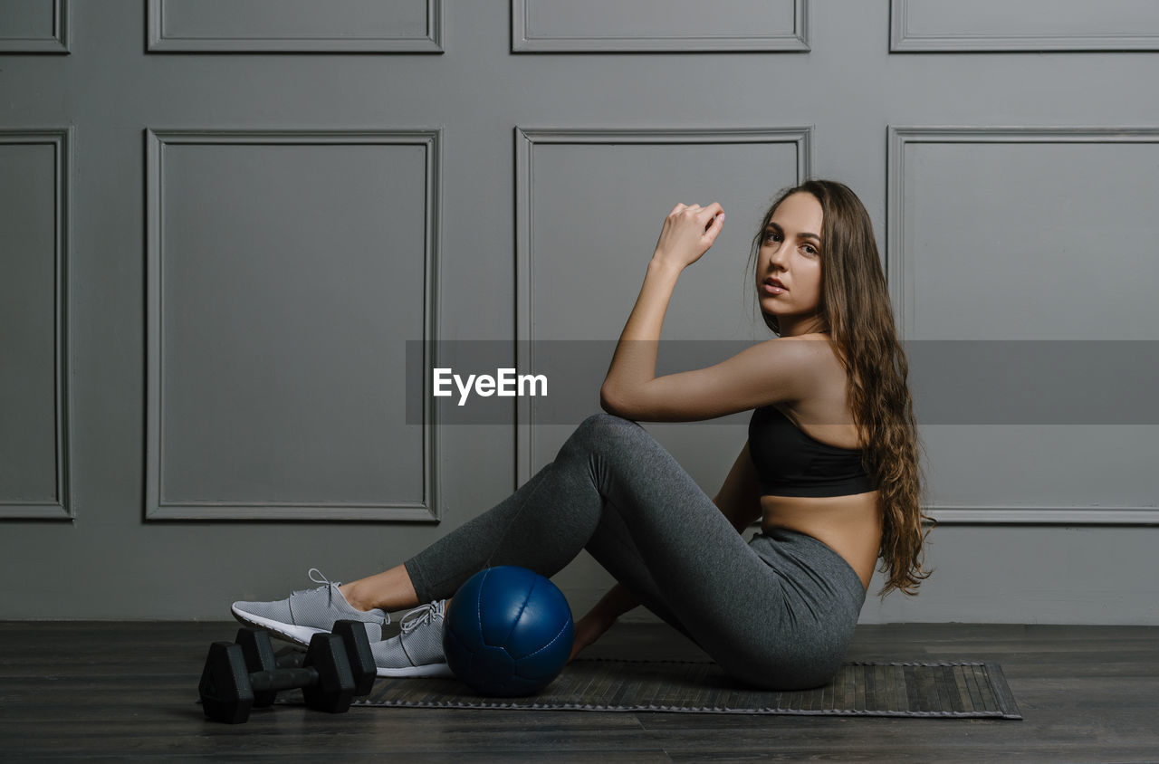 Portrait of young woman sitting by medicine ball against wall