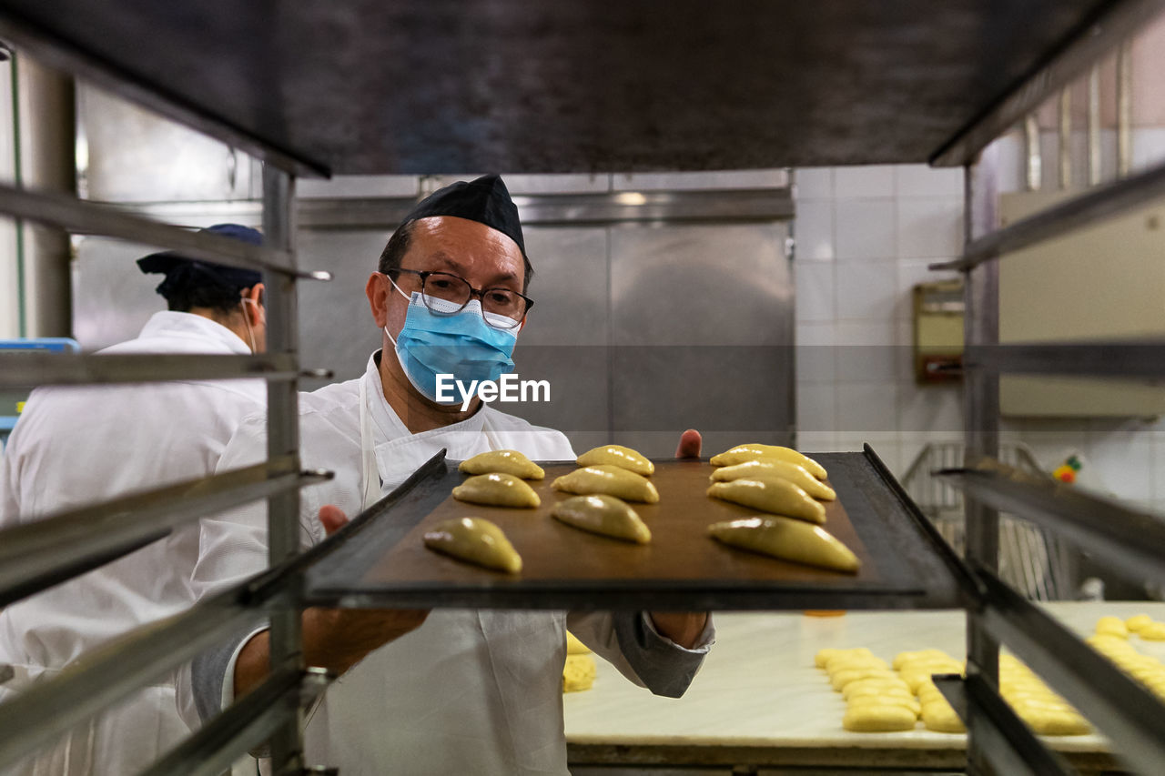 Male chef wearing uniform and medical mask preparing tasty pastry in kitchen of bakery during coronavirus epidemic