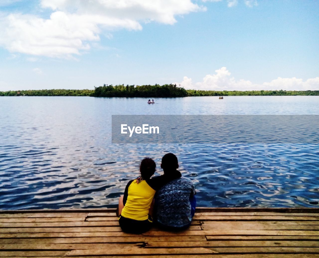 Rear view of couple sitting on pier by lake against sky
