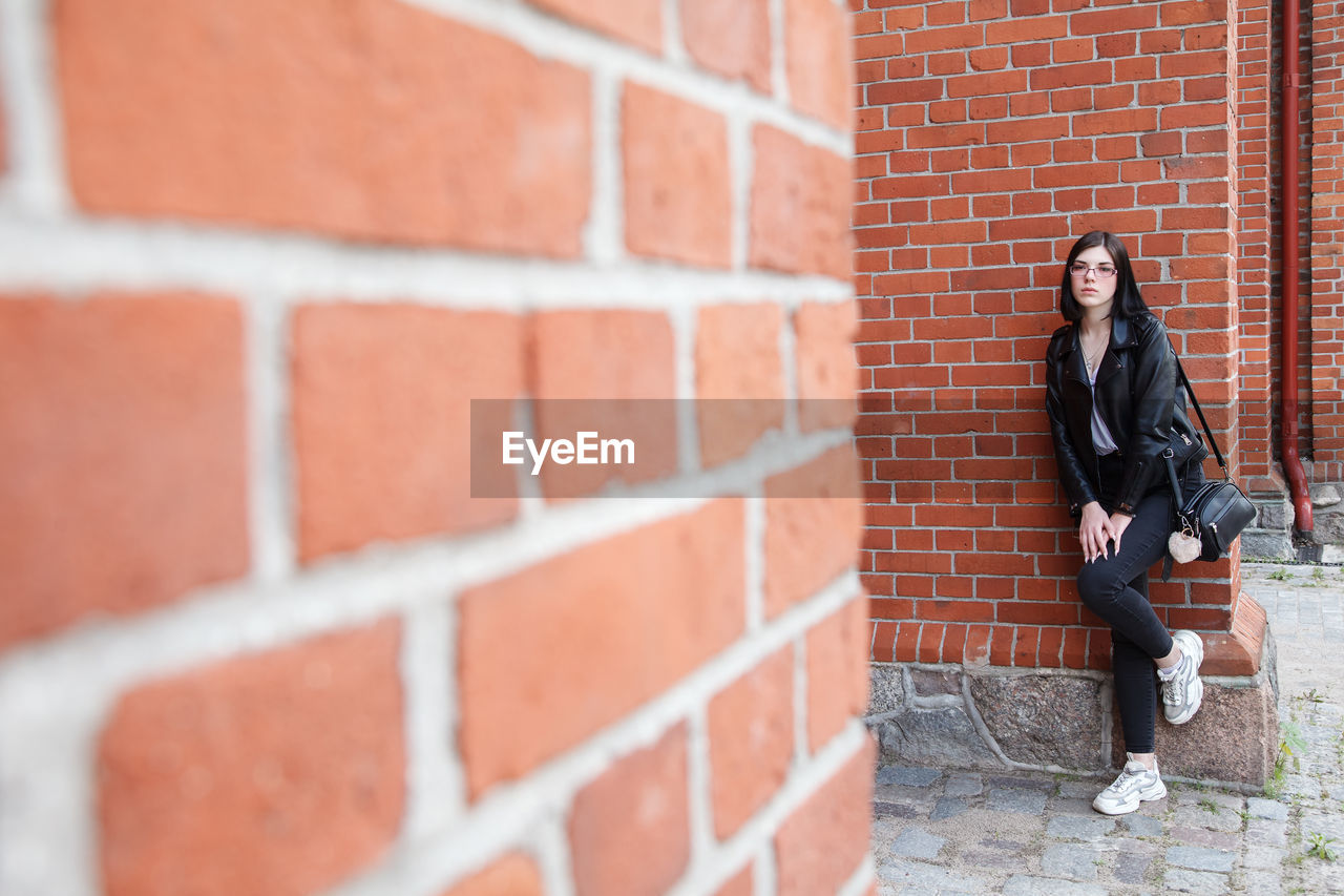 Young beautiful brunette girl in black jacket and jeans stands near wall of gothic church on summer