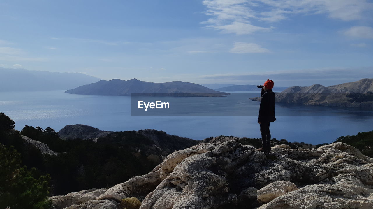 Boy looking through binoculars while standing on rocks by sea against sky