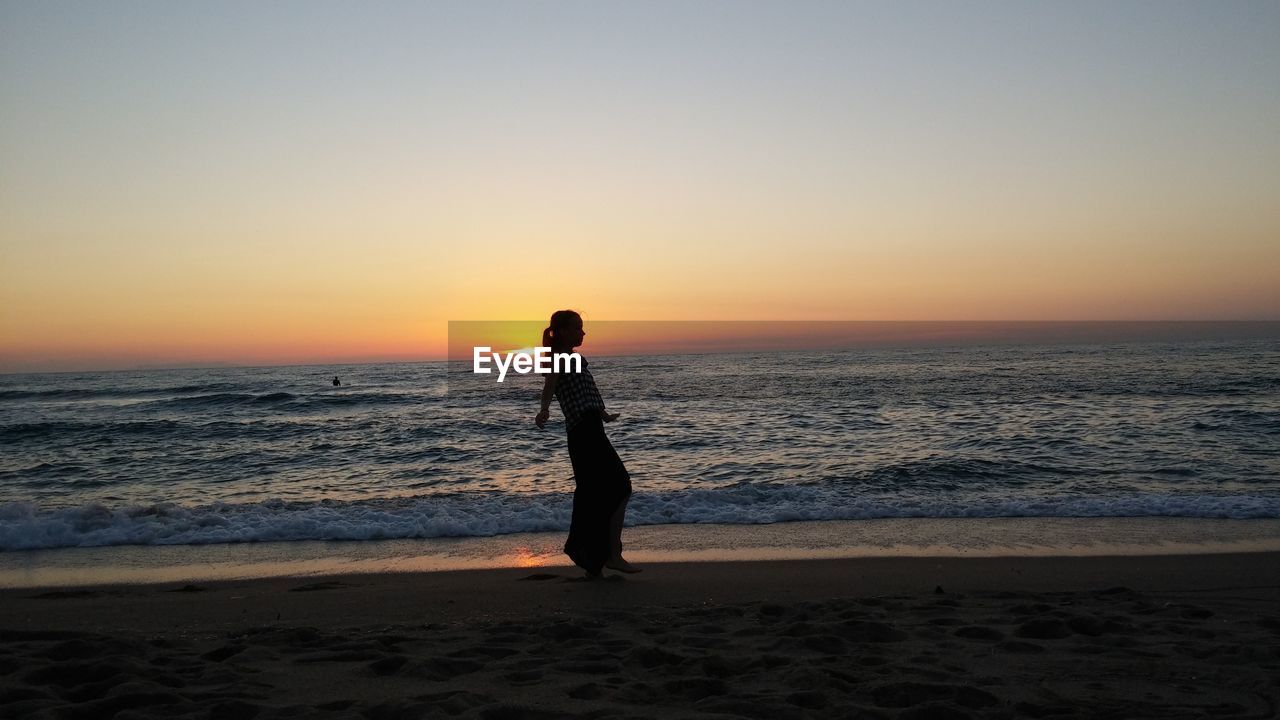 Woman walking at beach against sky during sunset