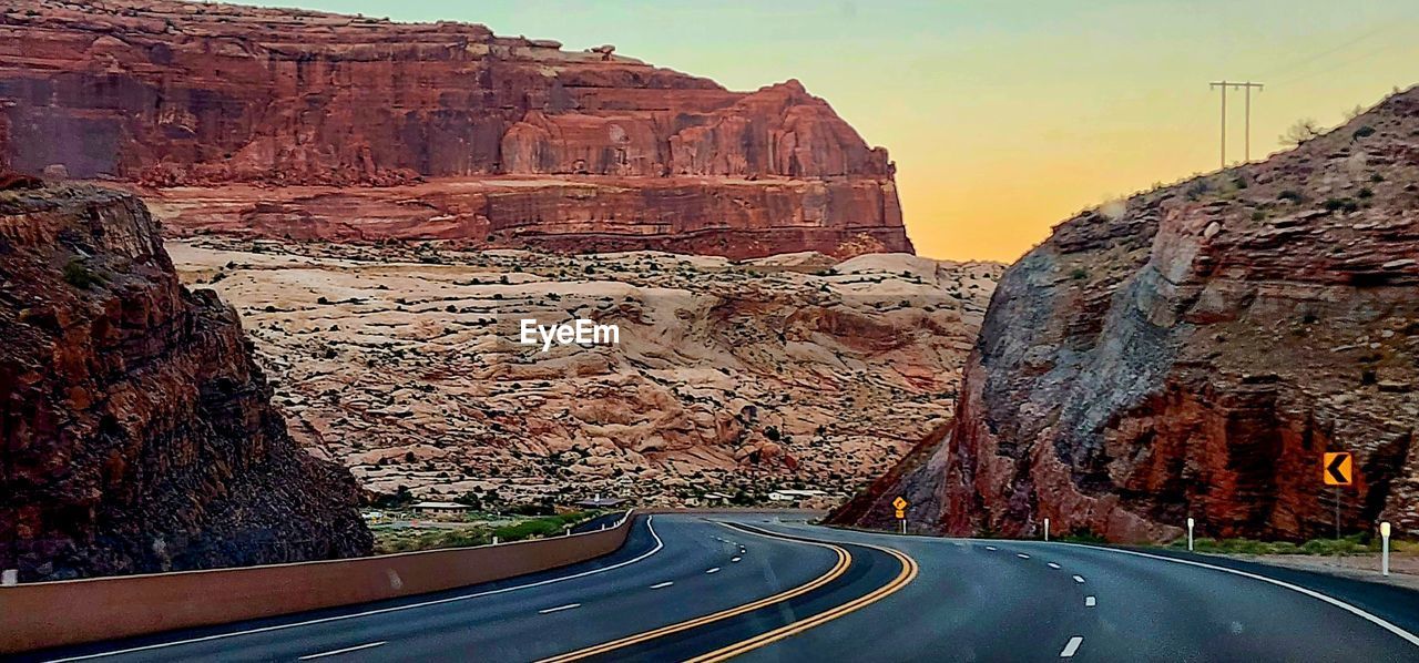 road, transportation, rock, nature, rock formation, canyon, symbol, travel destinations, travel, no people, sign, scenics - nature, sky, mountain, beauty in nature, non-urban scene, the way forward, landscape, arch, marking, infrastructure, road marking, environment, highway, land, curve, terrain, outdoors, cliff, mode of transportation, road trip, valley, street, wadi, tranquil scene, tranquility, empty road, sunset, desert, double yellow line, motor vehicle, escarpment