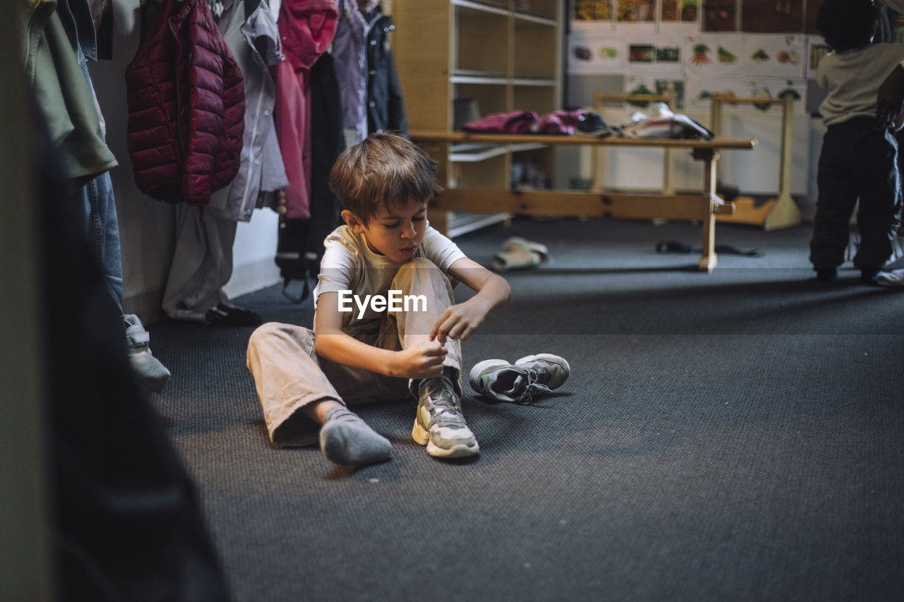 Boy tying shoelace while sitting on floor in cloakroom at preschool