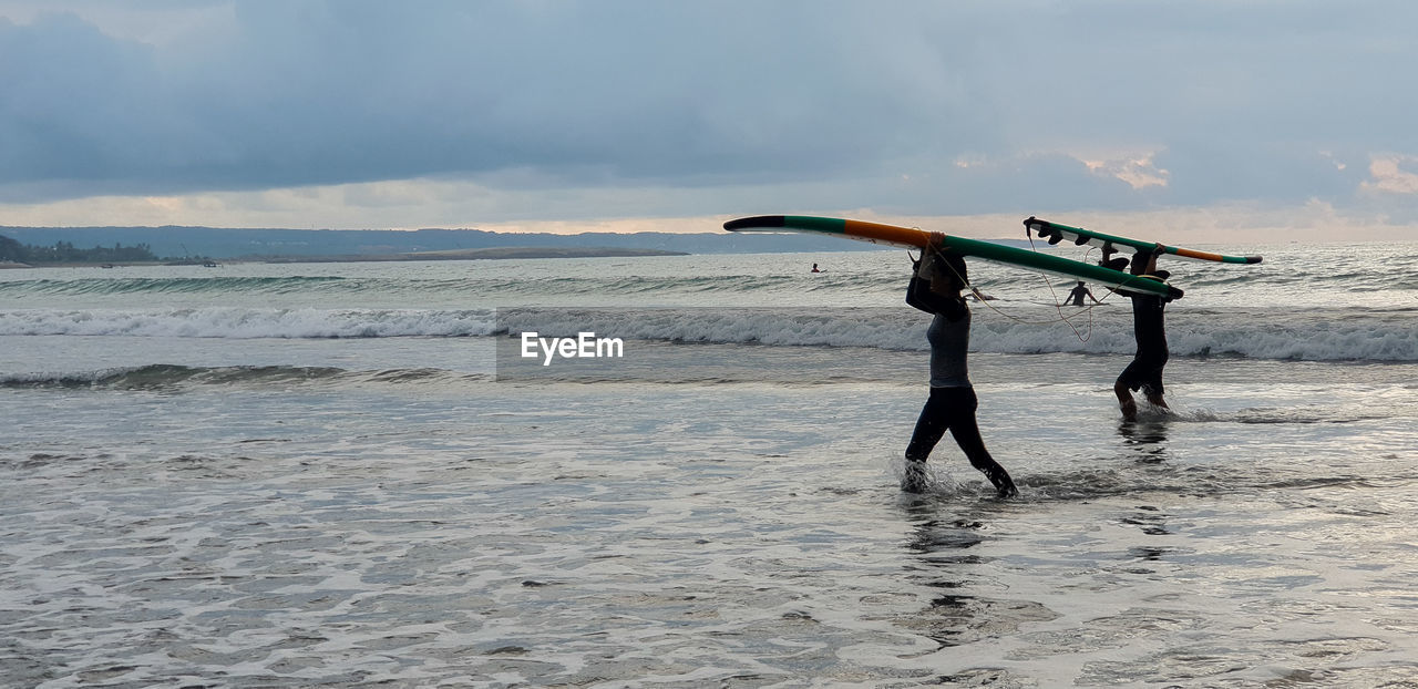 Side view of friends carrying surfboards on heads while wading at beach against sky during sunset