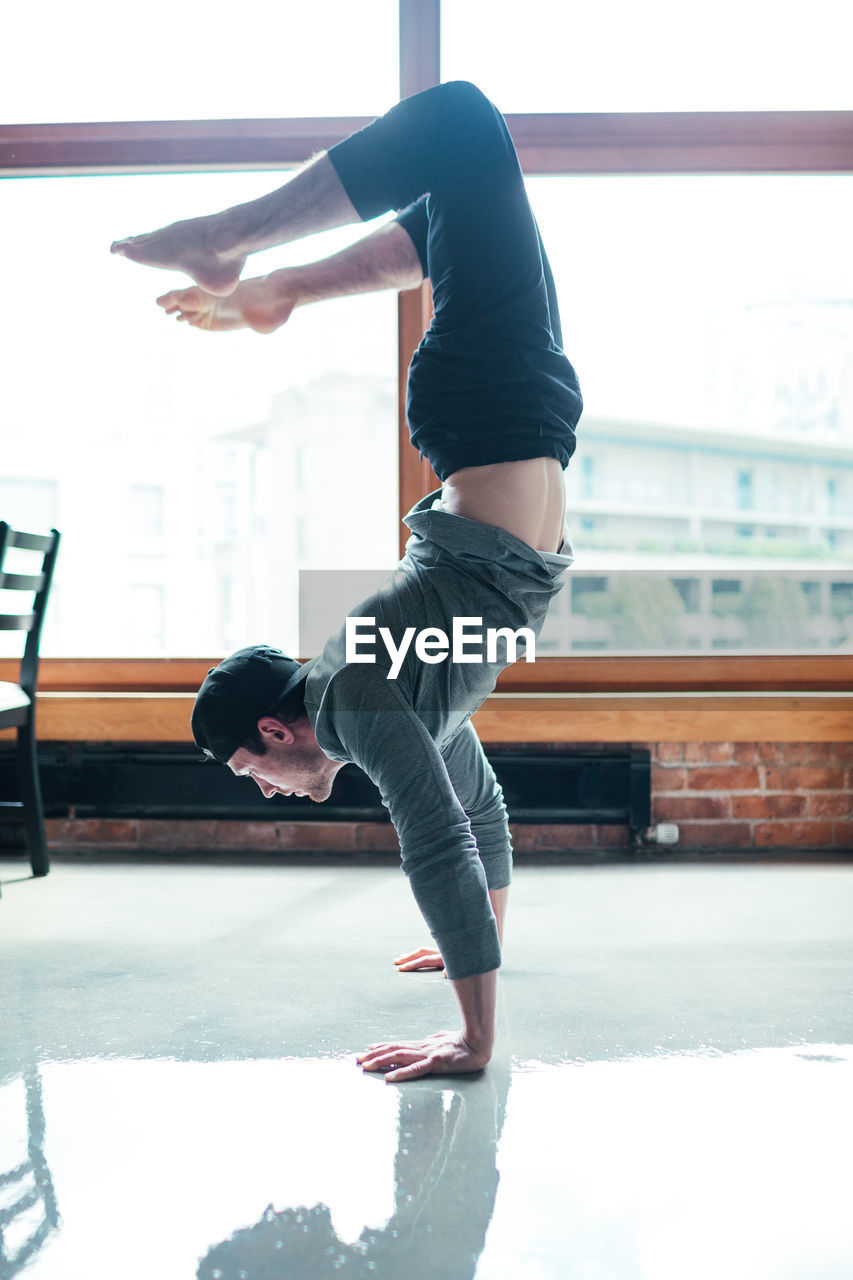 Side view of man practicing handstand on floor against window