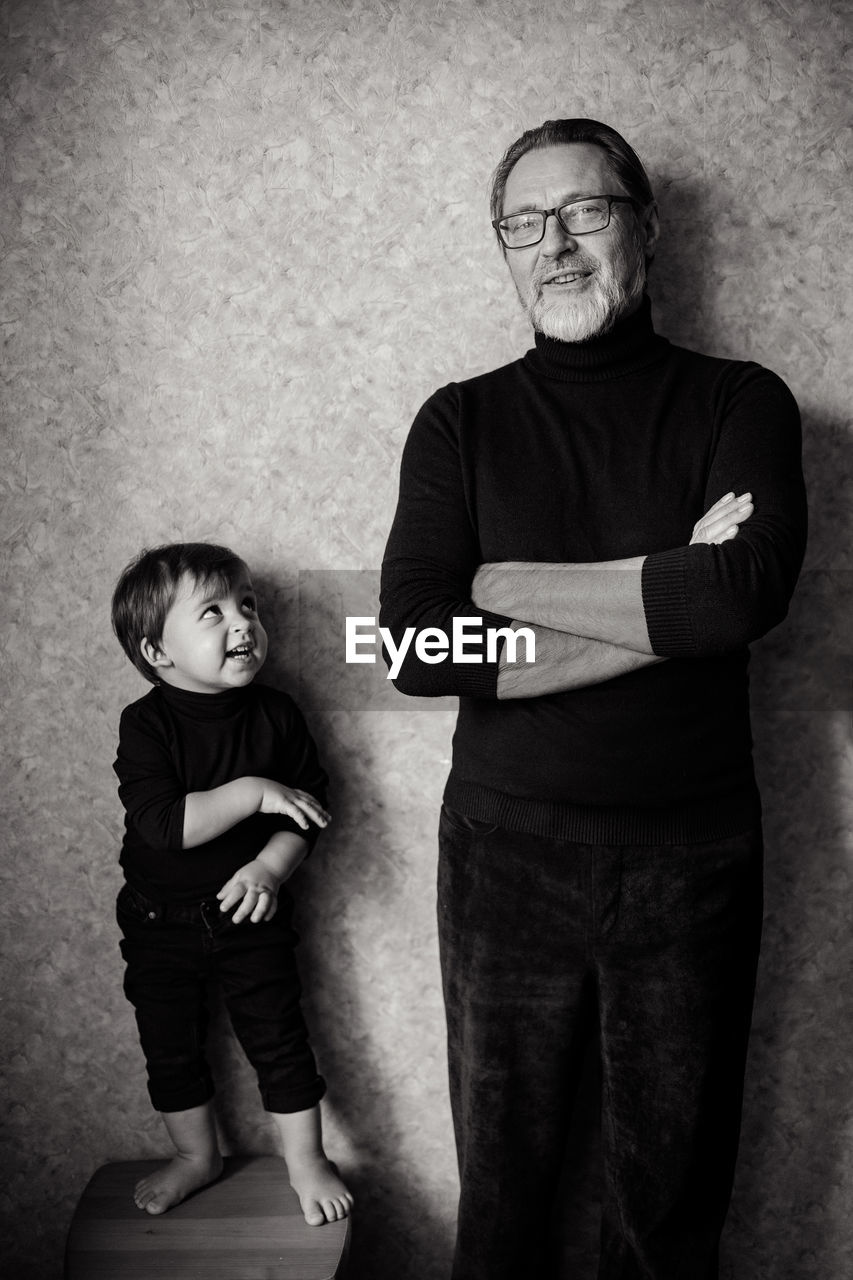 Grandfather with beard and glasses stands with his grandson in the studio near the wall