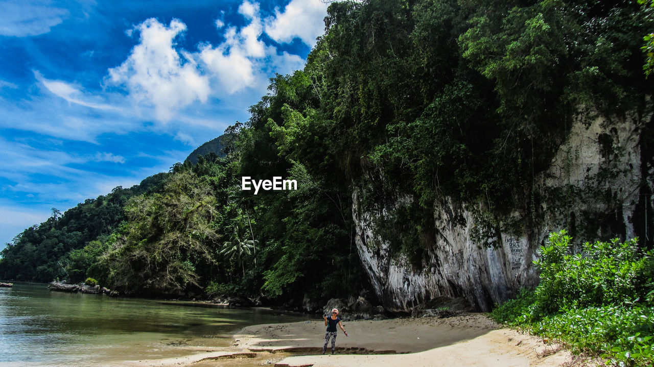 Woman at beach by rock formations and trees against cloudy sky