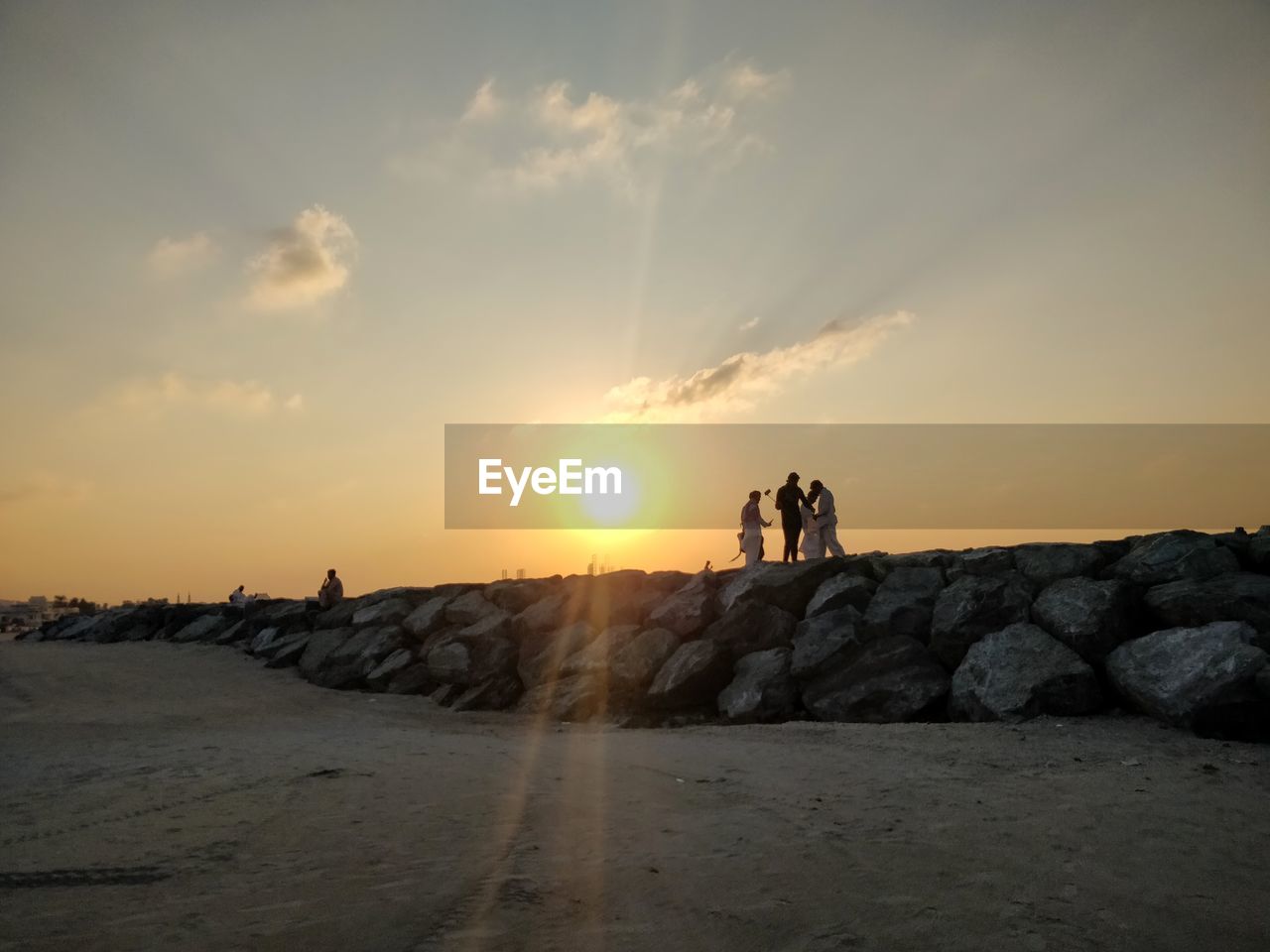 PEOPLE STANDING ON ROCKS BY LAND AGAINST SKY DURING SUNSET