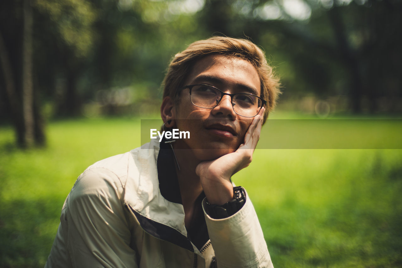 Thoughtful young man with hand on chin while sitting against trees in park