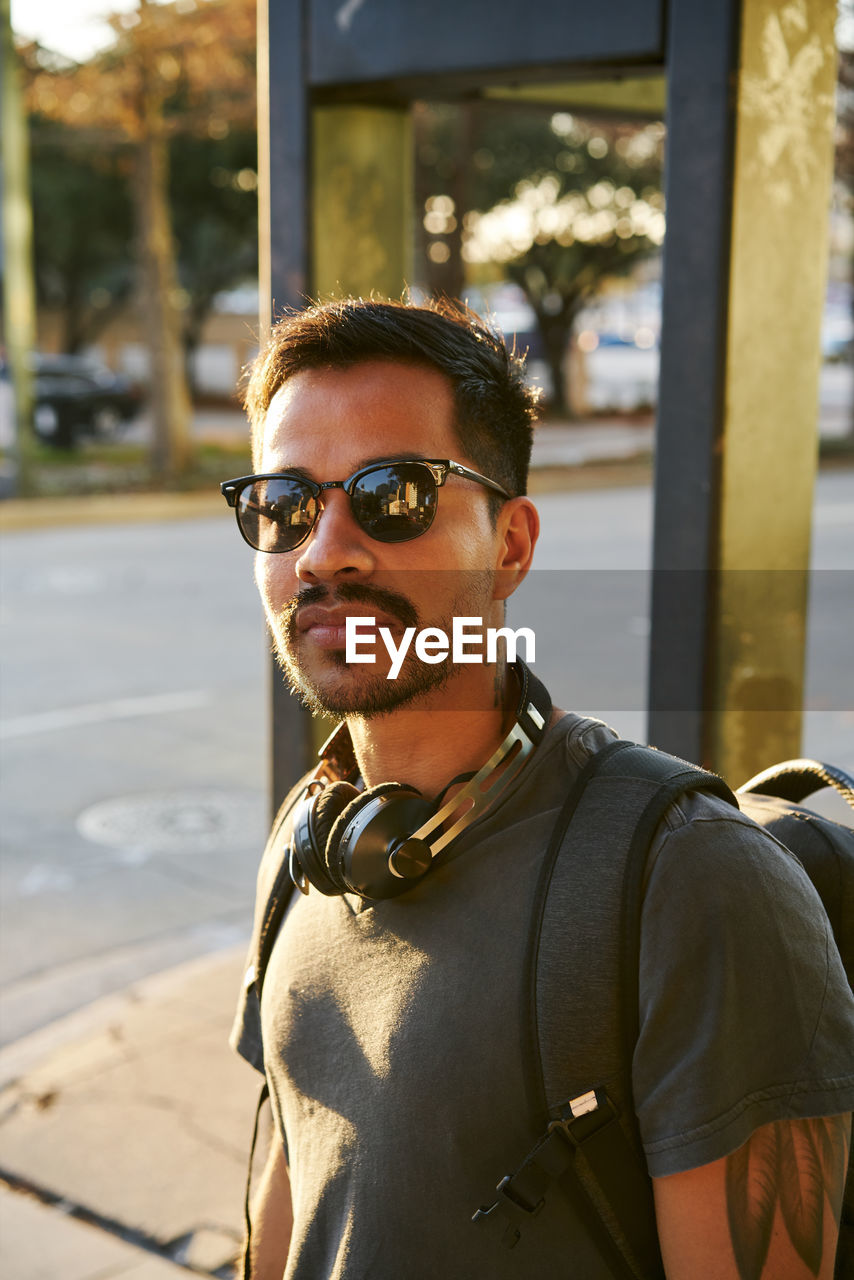 Young hispanic man in casual wear and stylish sunglasses with headphones and backpack looking at camera at city street