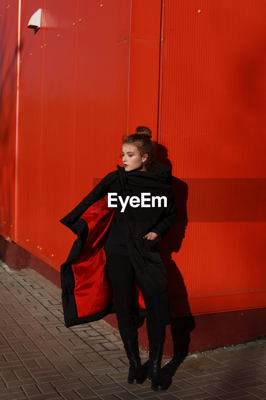 Fashionable teenage girl wearing black warm clothing standing against red wall during sunny day