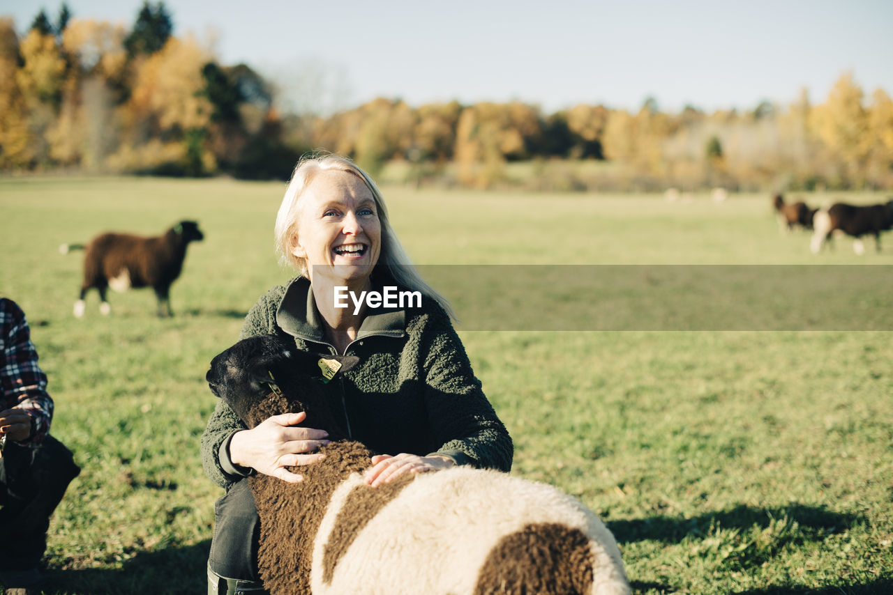 Smiling mature female farmer embracing sheep on field