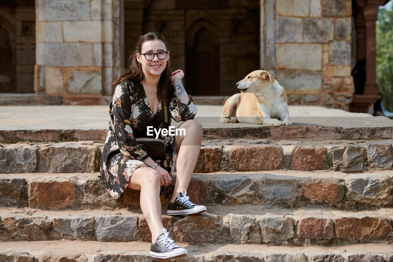 architecture, animal themes, sitting, animal, mammal, dog, adult, women, one animal, one person, smiling, happiness, domestic animals, pet, staircase, canine, portrait, spring, full length, young adult, fashion, emotion, built structure, glasses, hairstyle, long hair, wall, lifestyles, friendship, casual clothing, leisure activity, front view, day, clothing, person, looking at camera, cheerful, female, relaxation, building exterior, photo shoot, nature, outdoors, enjoyment, sunglasses, steps and staircases, positive emotion, city, carnivore, dress, brown hair, teeth, tourism, brick, city life
