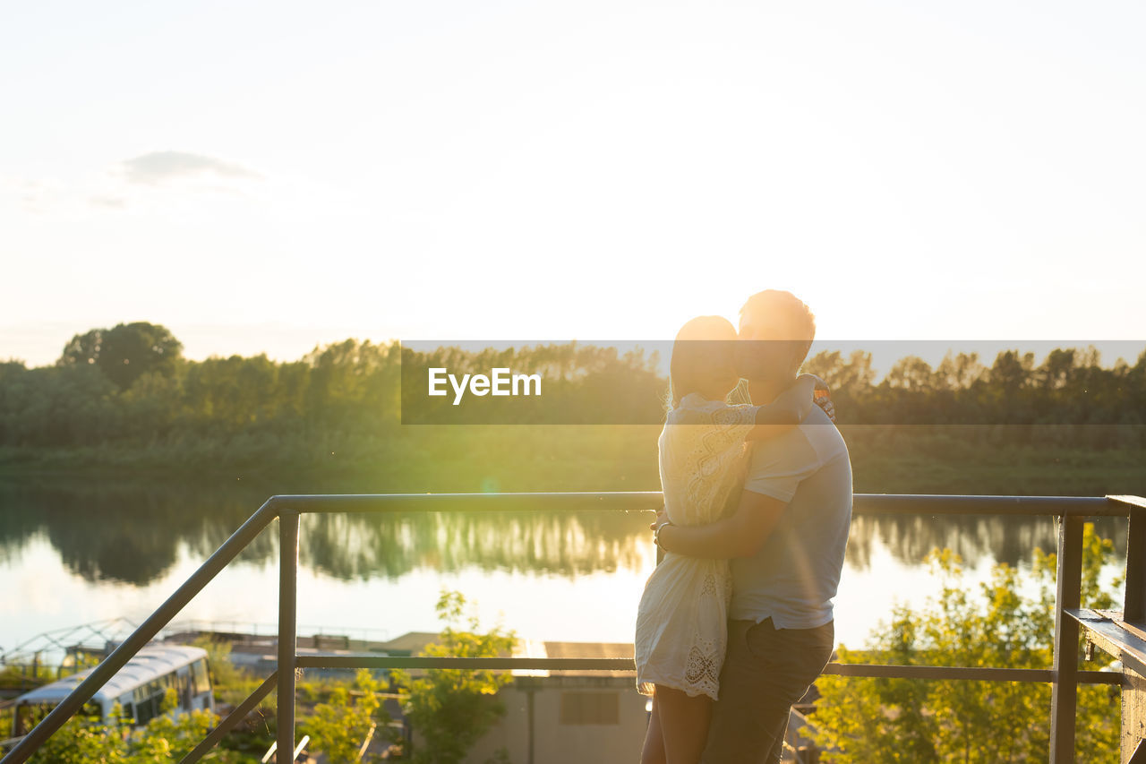 REAR VIEW OF COUPLE STANDING AT LAKE AGAINST SKY