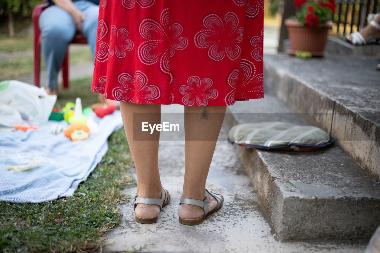 LOW SECTION OF WOMAN STANDING ON FOOTPATH BY RED PLANTS