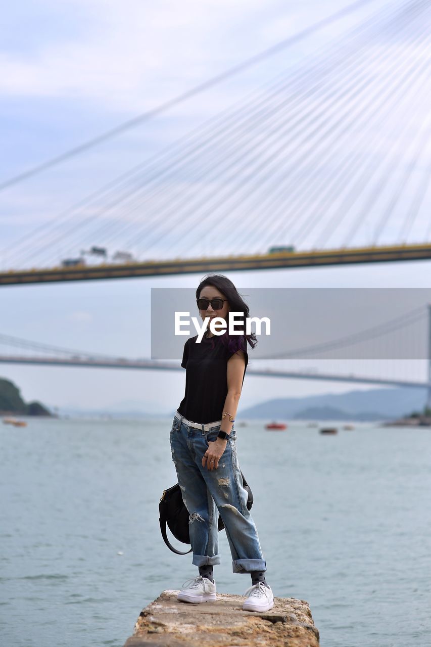 Full length of woman standing on rock against sea and bridge