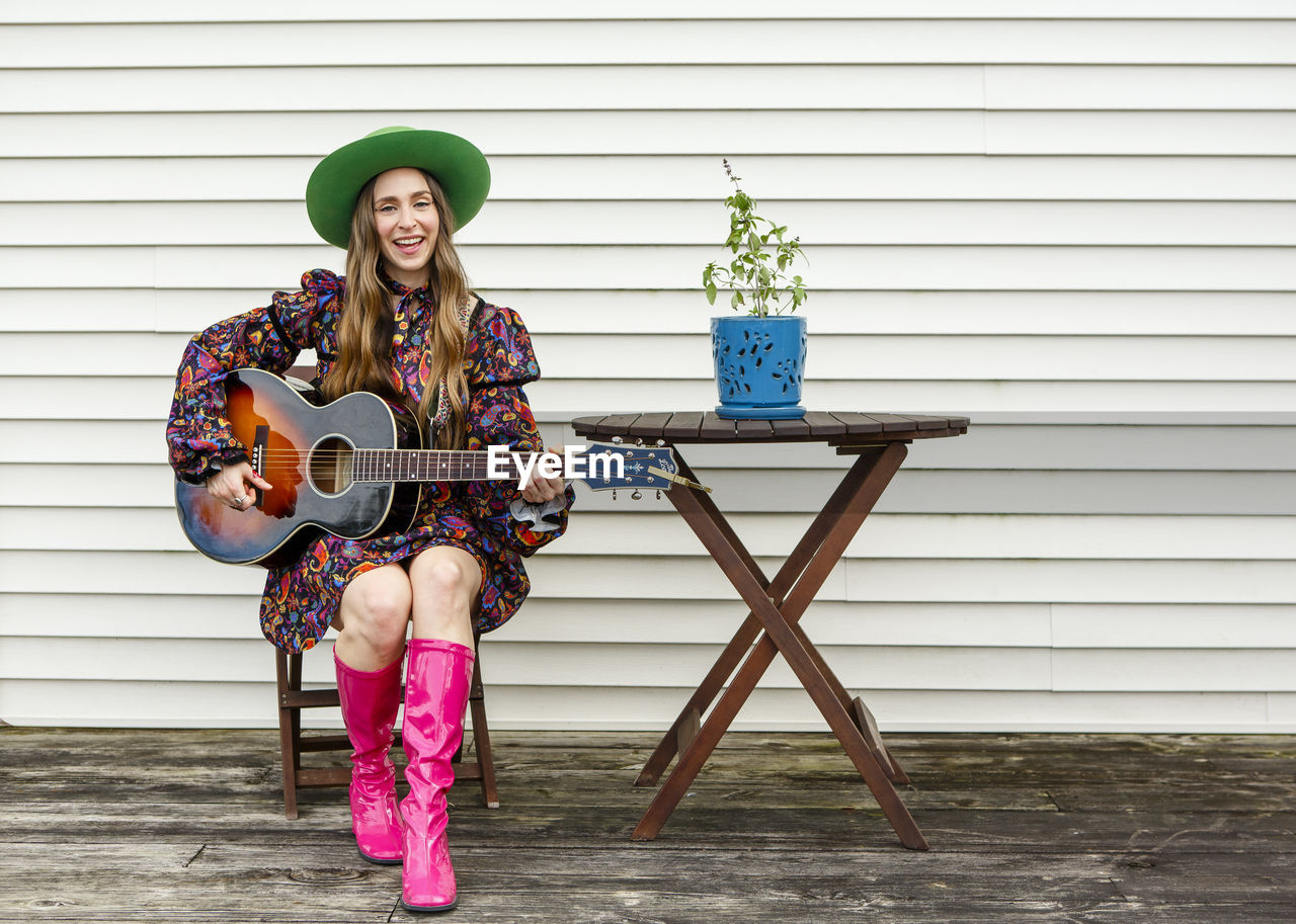 A woman sits alone at a wooden table on deck playing acoustic guitar