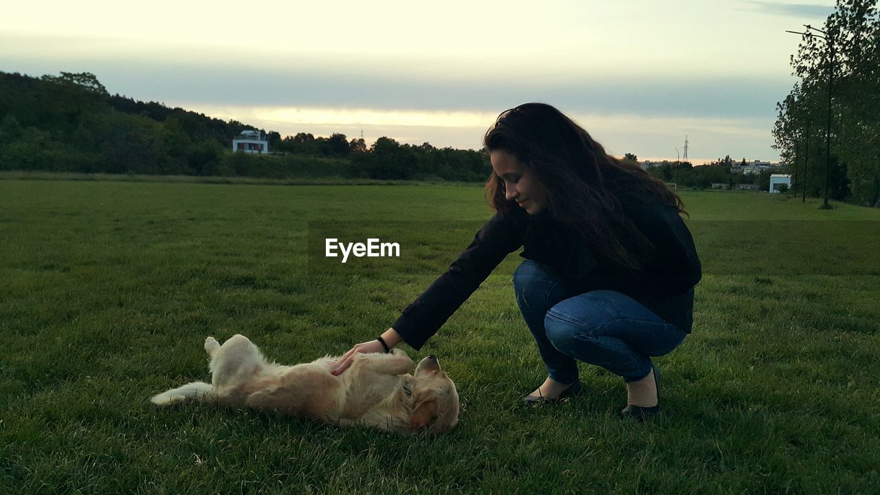 Full length of smiling woman playing with dog while crouching on grassy field