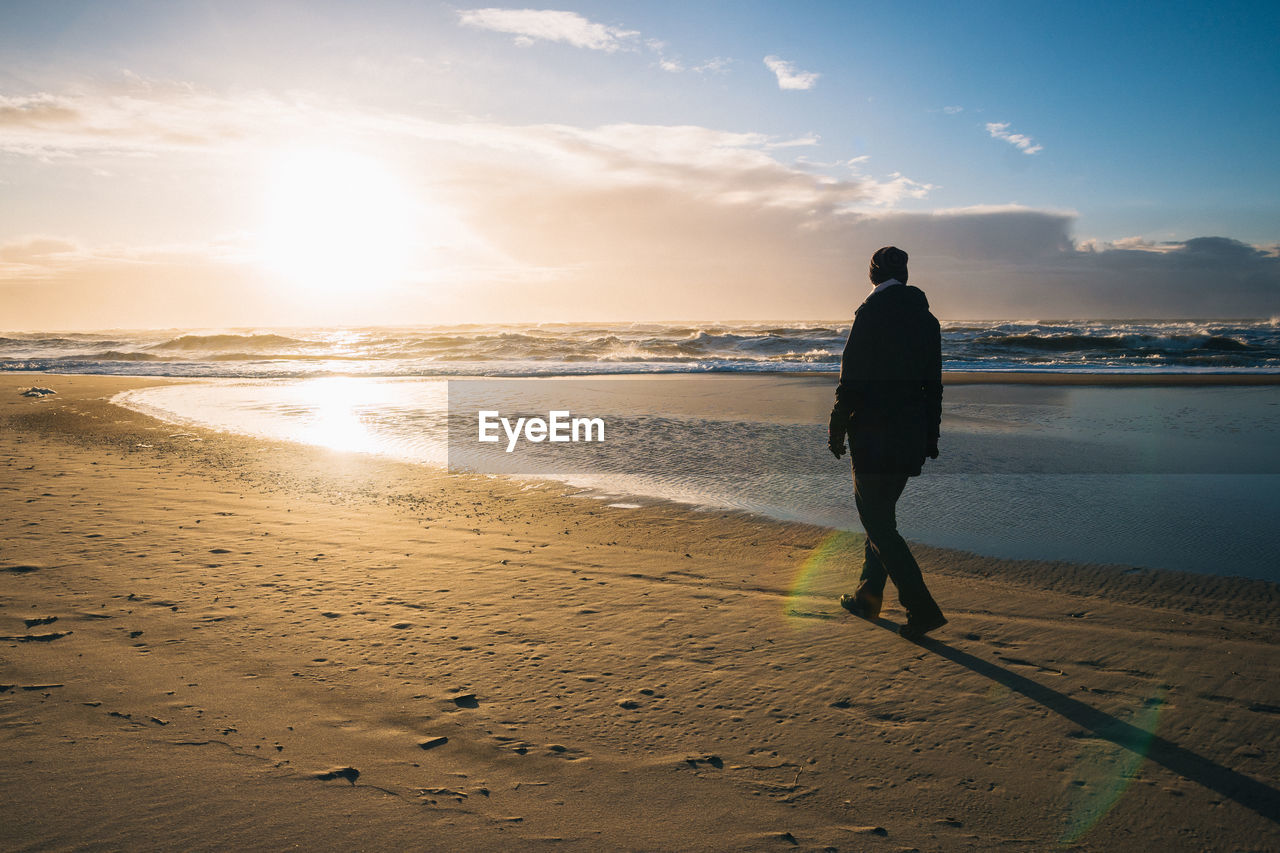 Man walking at beach against sky during sunset