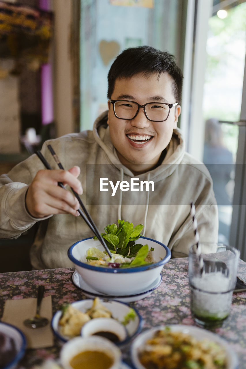 Portrait of smiling young man with food holding chopsticks at restaurant