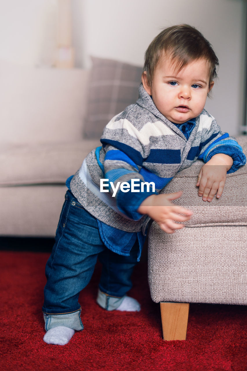 Portrait of cute baby boy standing by sofa at home
