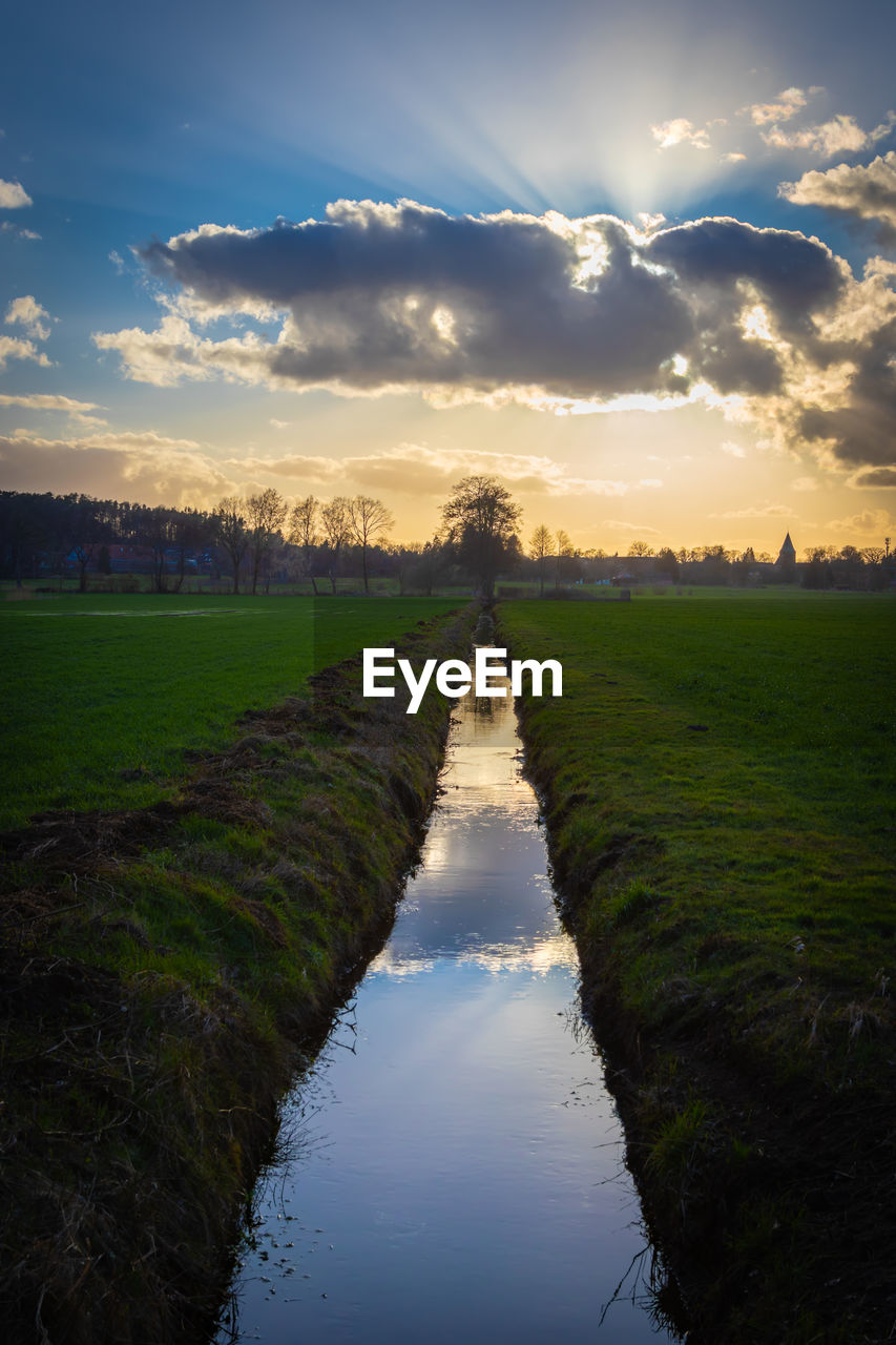 Canal amidst field against sky during sunset