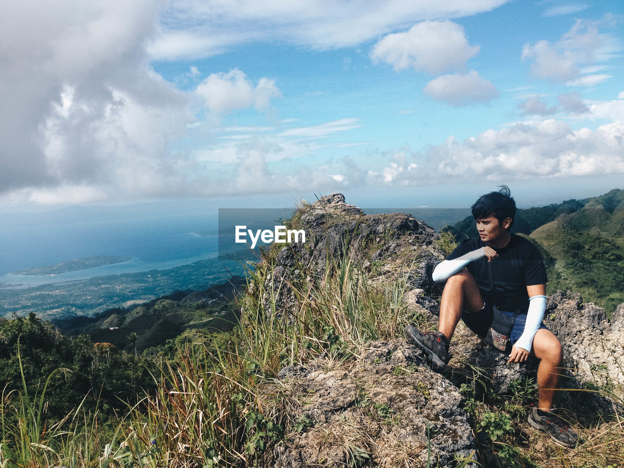 Young man sitting on mountain peak against cloudy sky