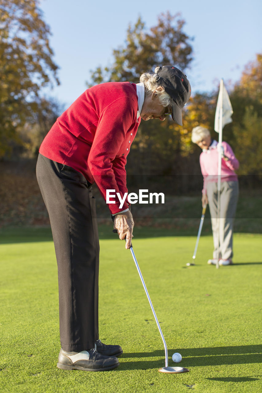 Side view of senior woman putting while friend holding flag on golf course
