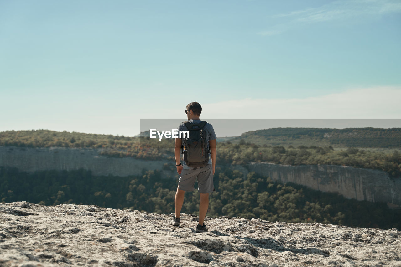 Back view of traveling man standing with backpack on rough hill and enjoying view of highland valley during summer holiday