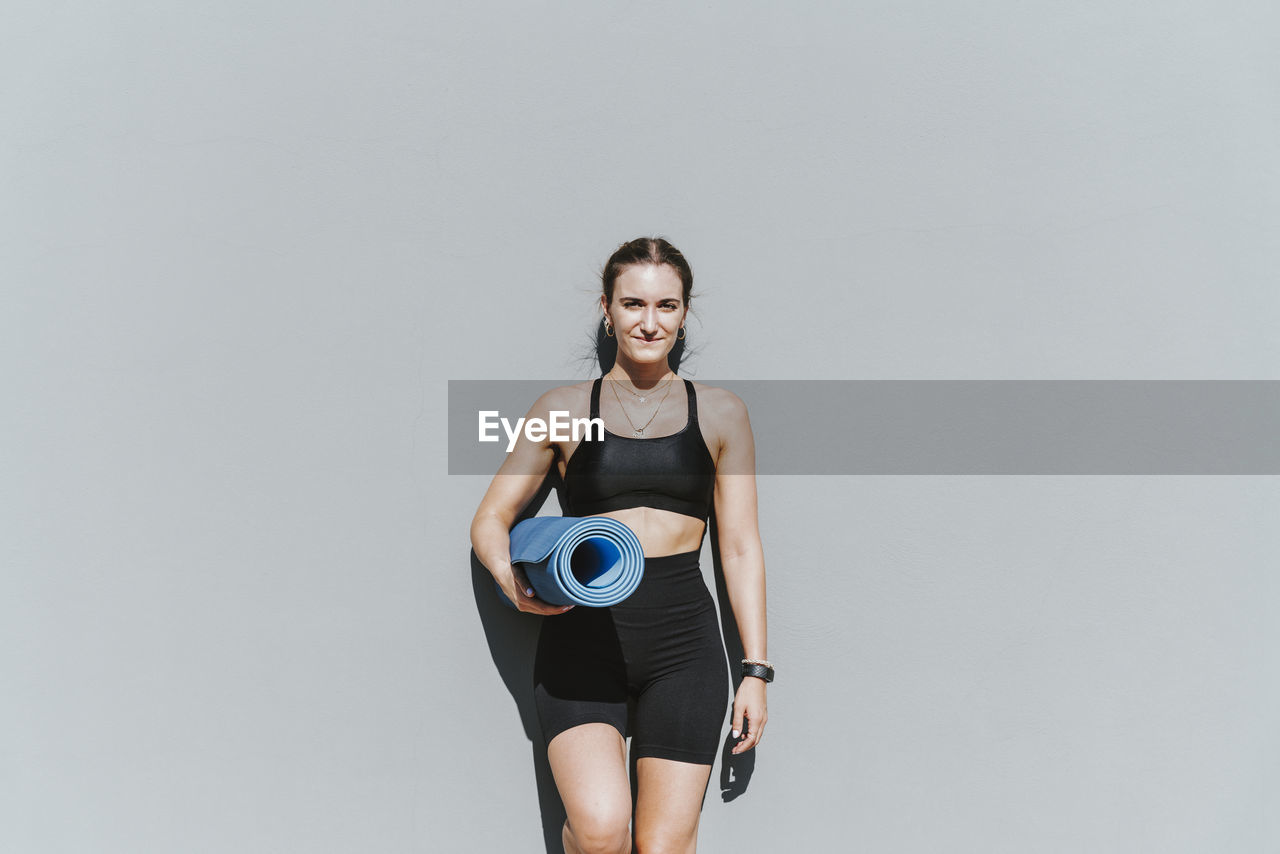 Smiling beautiful young woman with exercise mat leaning on wall