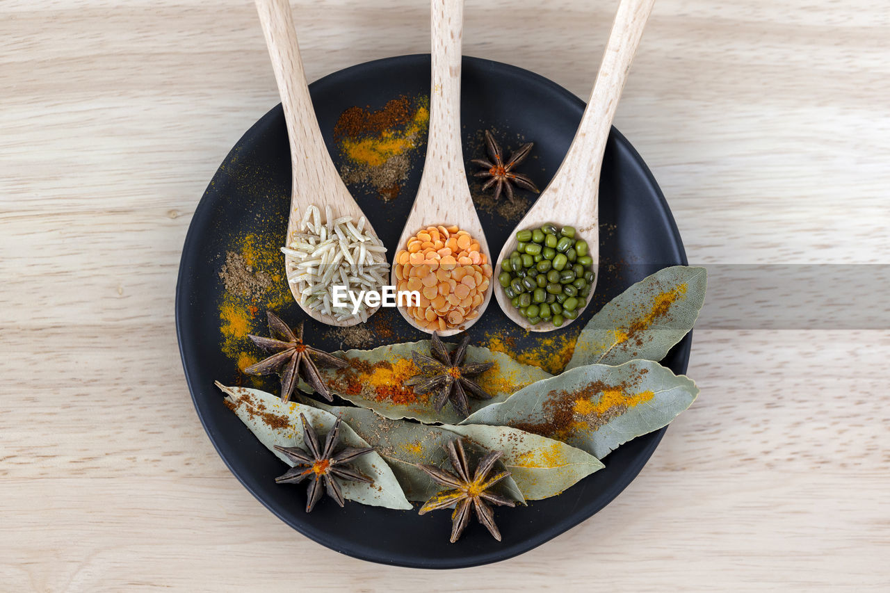 Rice, moong beans and dal in wooden spoons next to star anise seeds, bay leaf and colorful spices