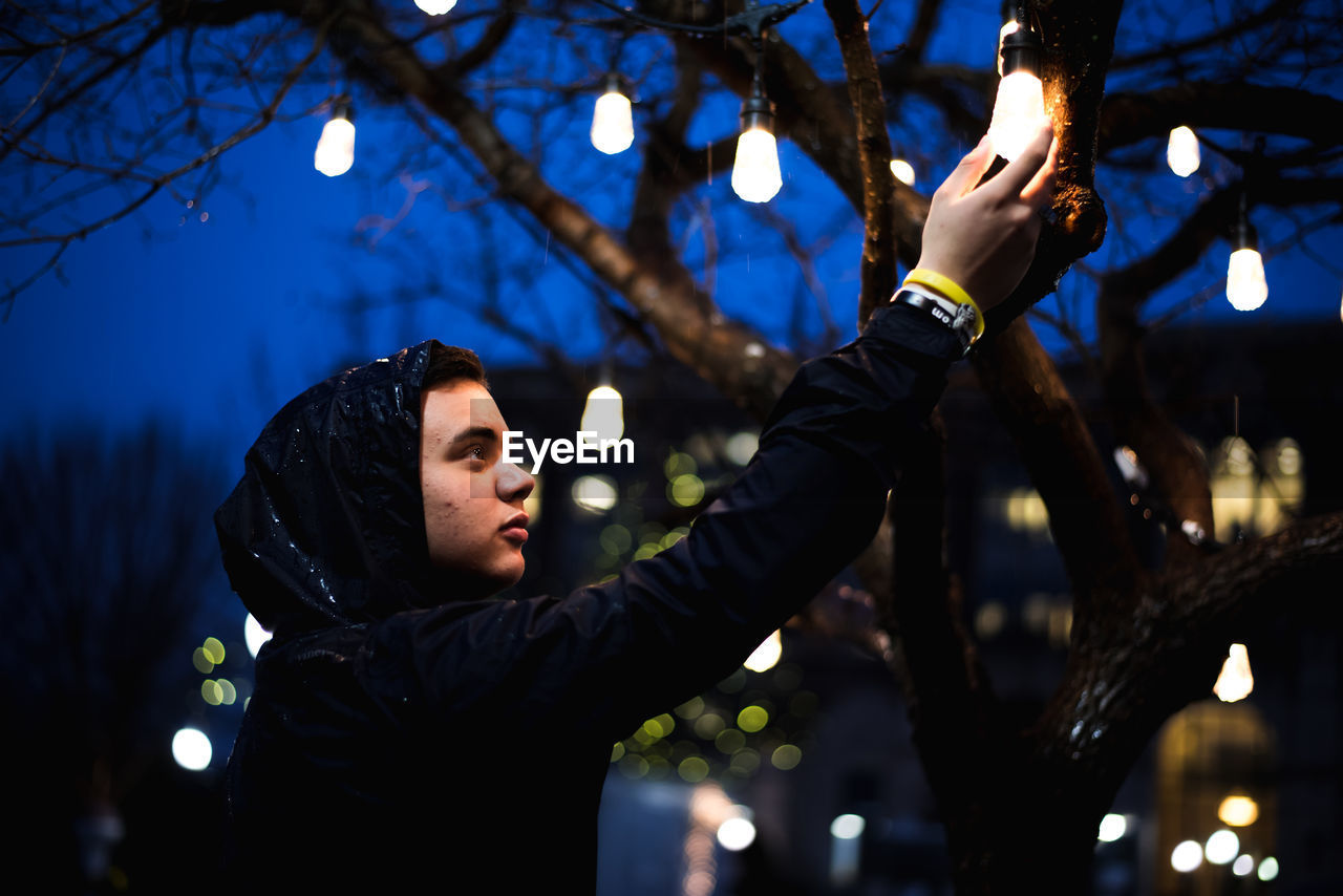 Low angle view of man holding illuminated light bulb hanging from tree at night