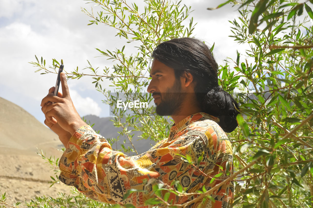 Side view of a long haired young guy taking photo with his phone with the background of mountains 