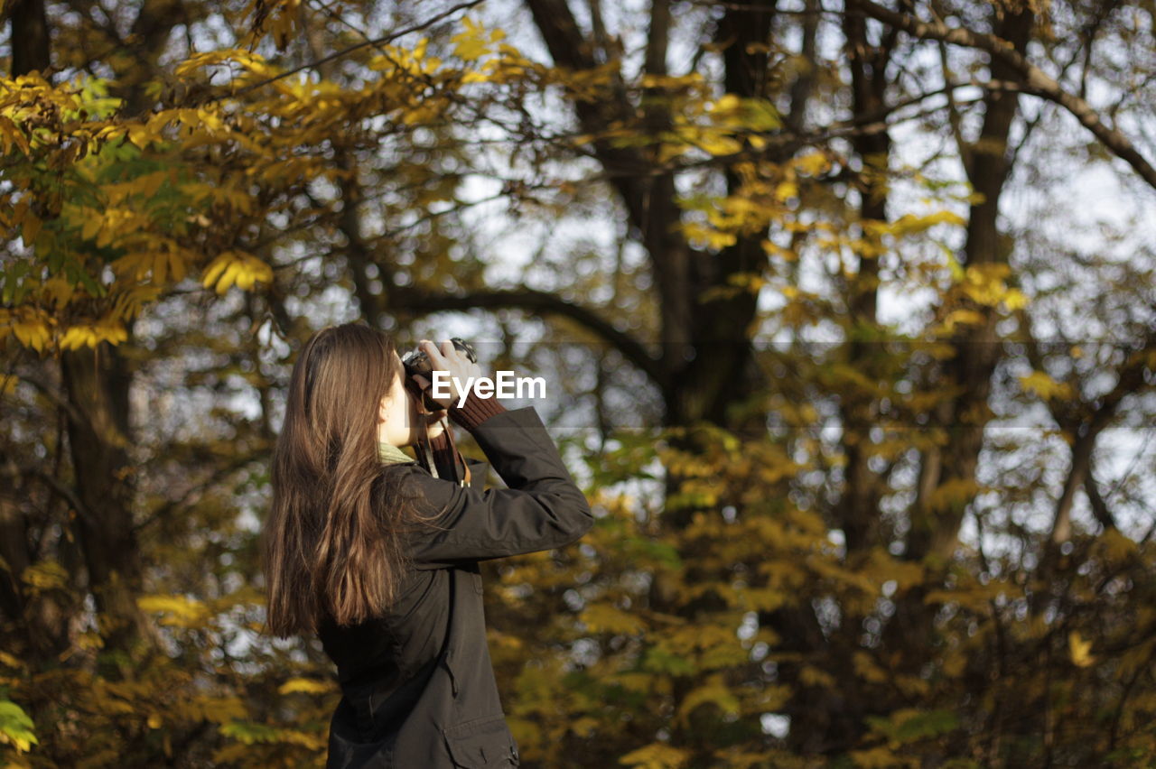 Side view of young woman photographing while standing against trees in forest