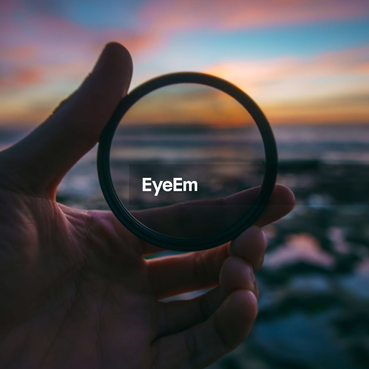 Cropped image of hand holding lens at beach during sunset