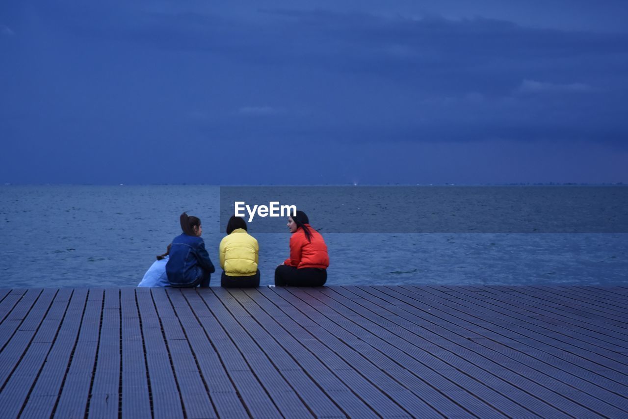 REAR VIEW OF PEOPLE SITTING ON SHORE AGAINST SEA