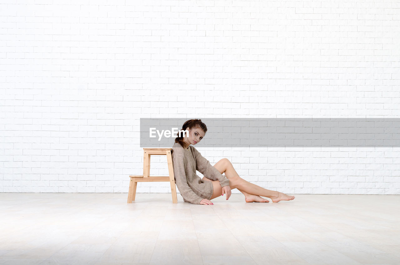 Full length portrait of young woman sitting by wood stool against brick wall
