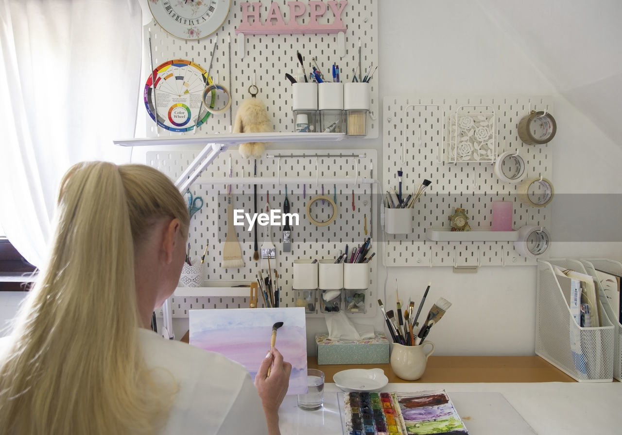 Back view of a blonde girl draws in a home studio equipped in modern design.