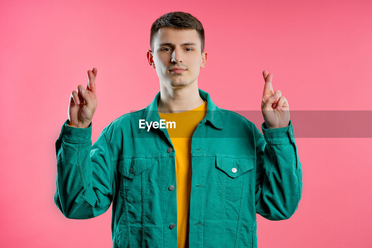 colored background, studio shot, one person, portrait, looking at camera, young adult, waist up, front view, sign language, adult, gesturing, outerwear, indoors, men, standing, emotion, pink background, hood, jacket, smiling, hand sign, pink, clothing, sleeve, casual clothing, individuality, happiness, green, person