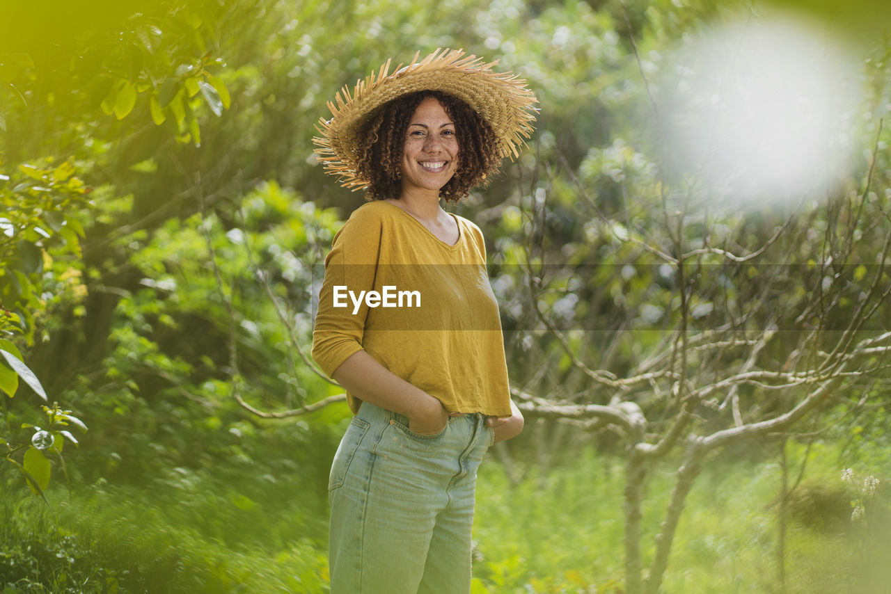 Smiling woman in straw hat standing by bare tree in vegetable garden